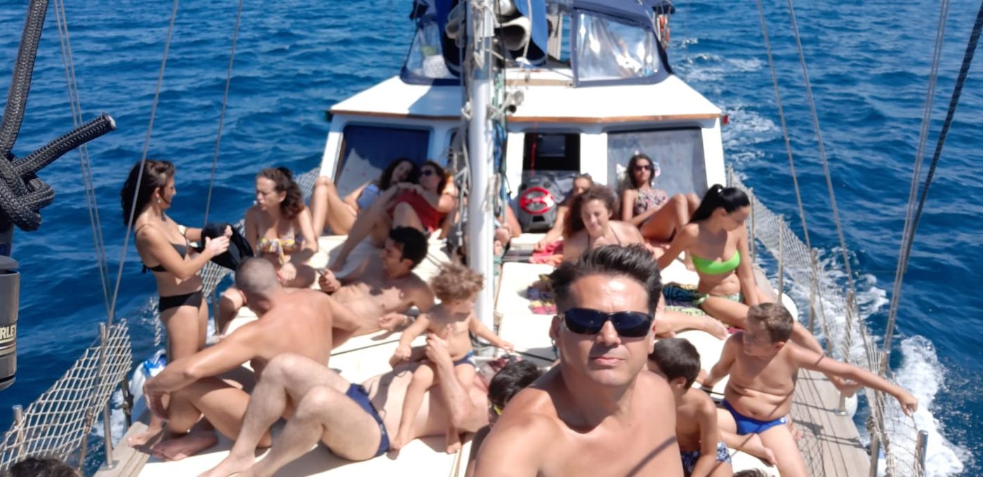 Gruppo di adulti e bambini in costume sul ponte di una barca a vela con reti laterali, in navigazione sul mare blu
